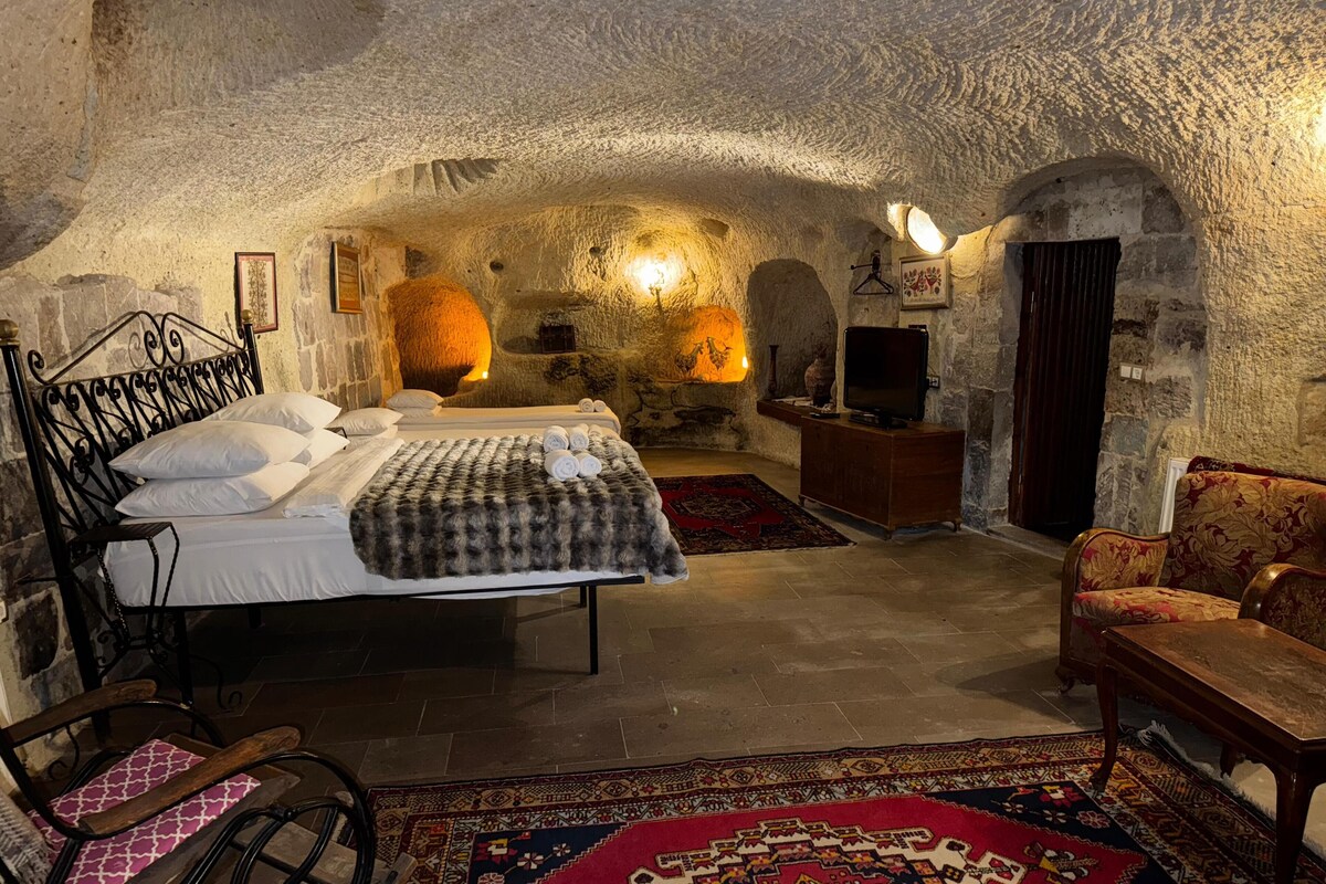 A rustic stone-walled bedroom with a large bed, a wooden chair, a television, and a fireplace.