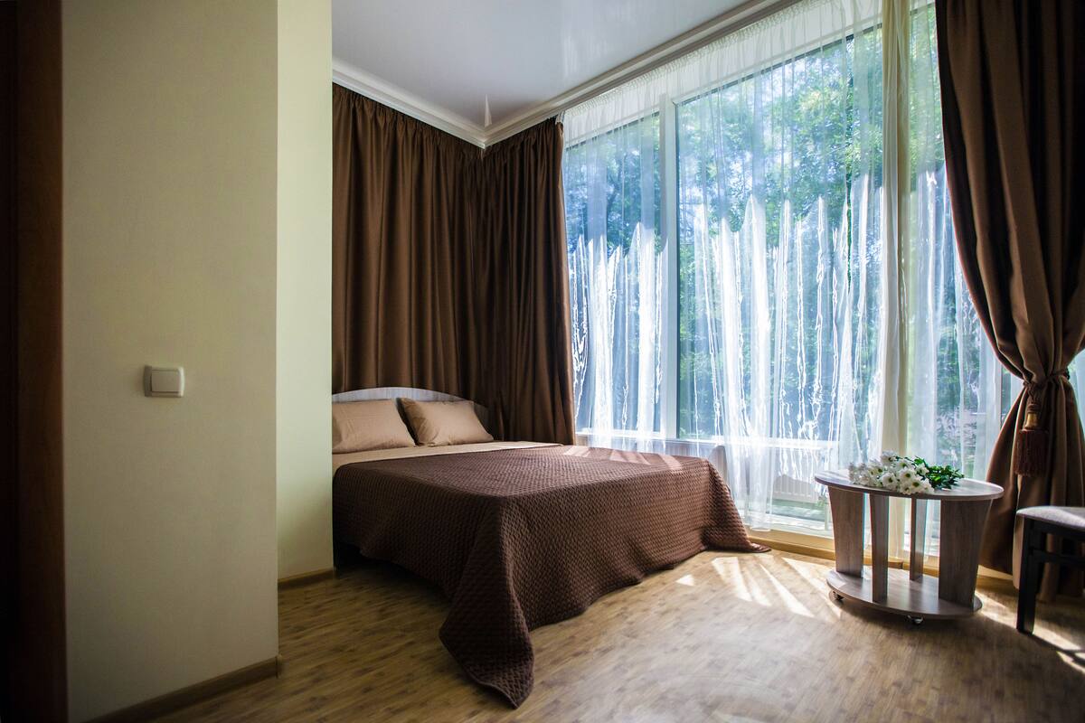 A bedroom with a large window, a bed with a brown bedspread, and a small round table with a plant.