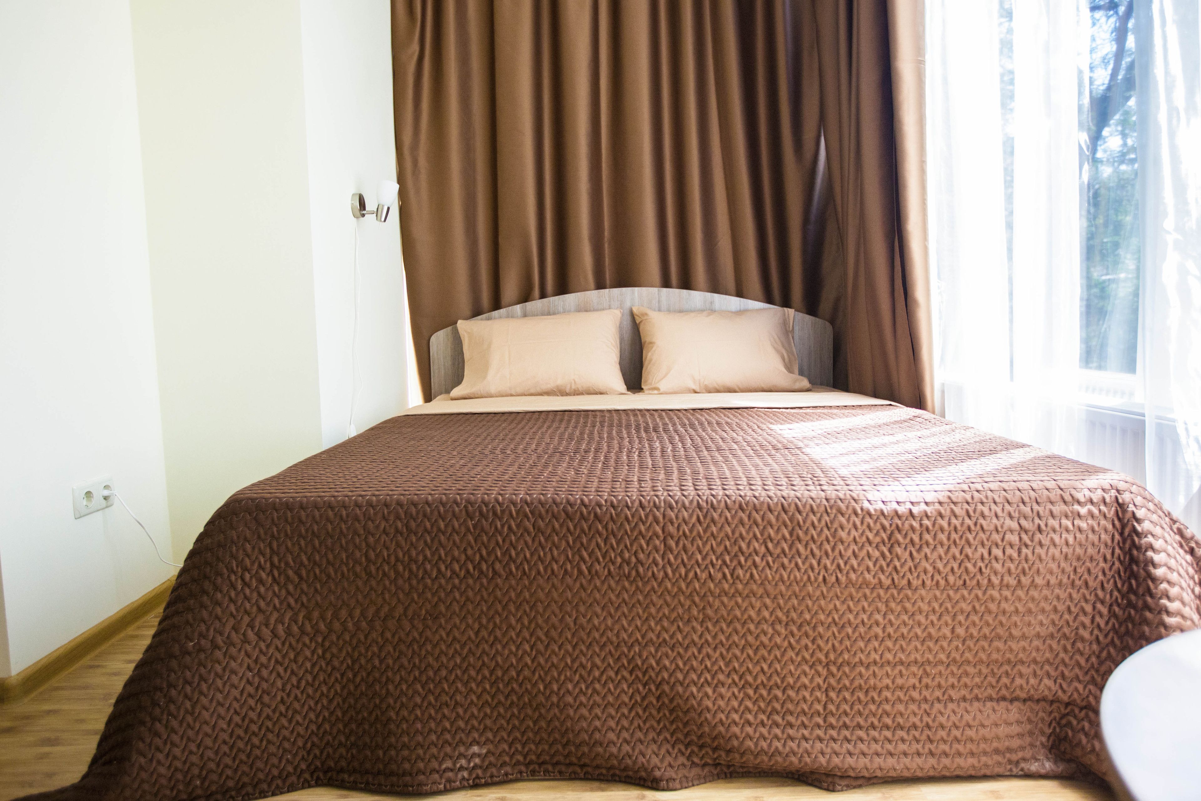 A neatly made bed with a brown textured bedspread and two pillows, a window with curtains, and a power outlet on the wall.
