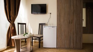 Una habitación compacta con piso de madera, una pequeña mesa redonda con una silla, un refrigerador blanco y un televisor instalado en la pared.