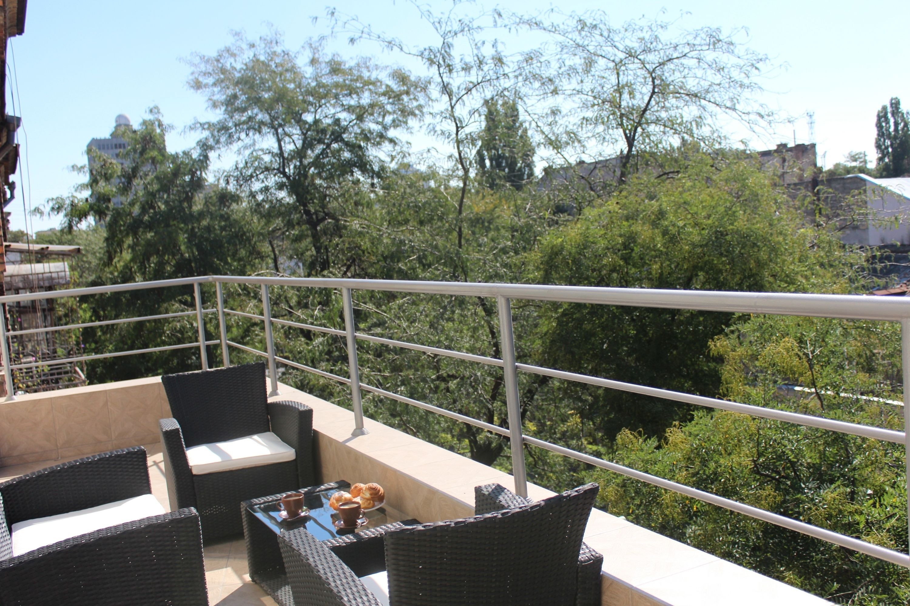Un balcón con mobiliario de mimbre y una mesa, con vista a un jardín con árboles y edificios al fondo.