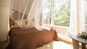 Un dormitorio con una cama cubierta por un edredón marrón, una mesita de noche con un jarrón de flores y una ventana que da a vistas de vegetación.