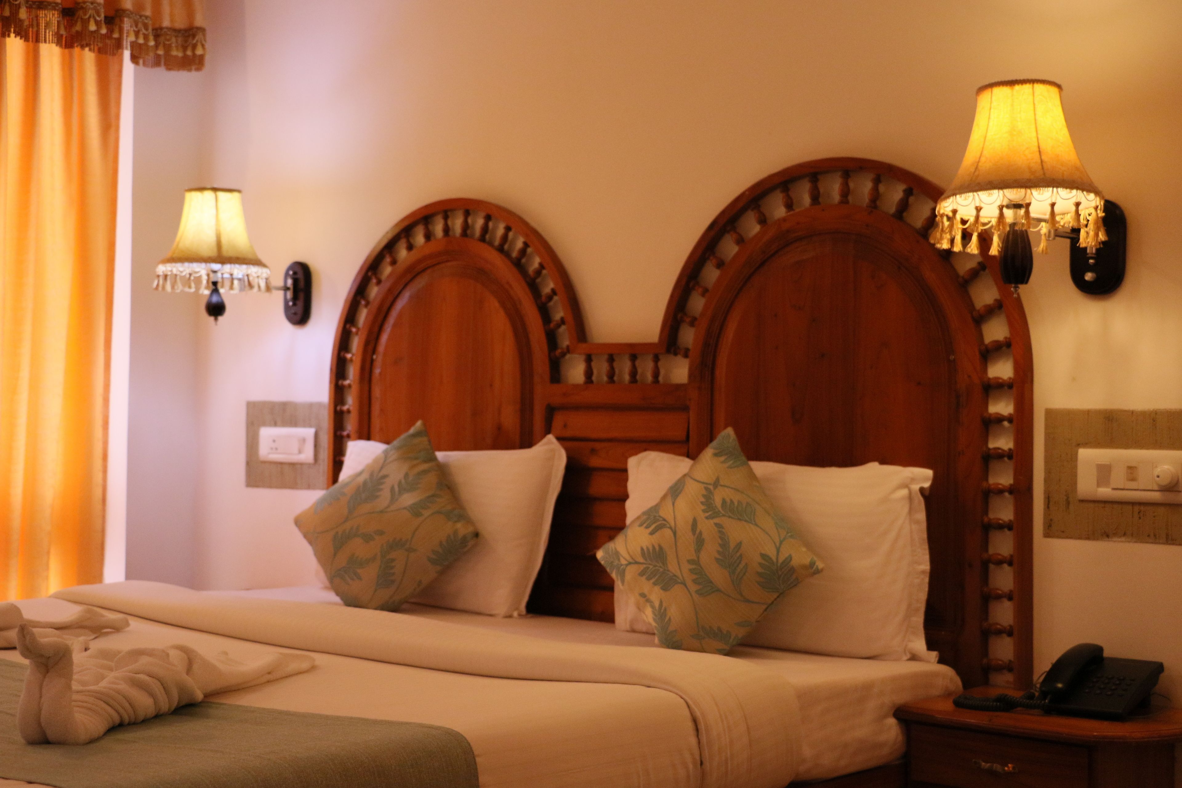 Habitación de hotel con cabecera de madera, dos lámparas de pared, una cama con ropa blanca y una mesita de noche con un teléfono.