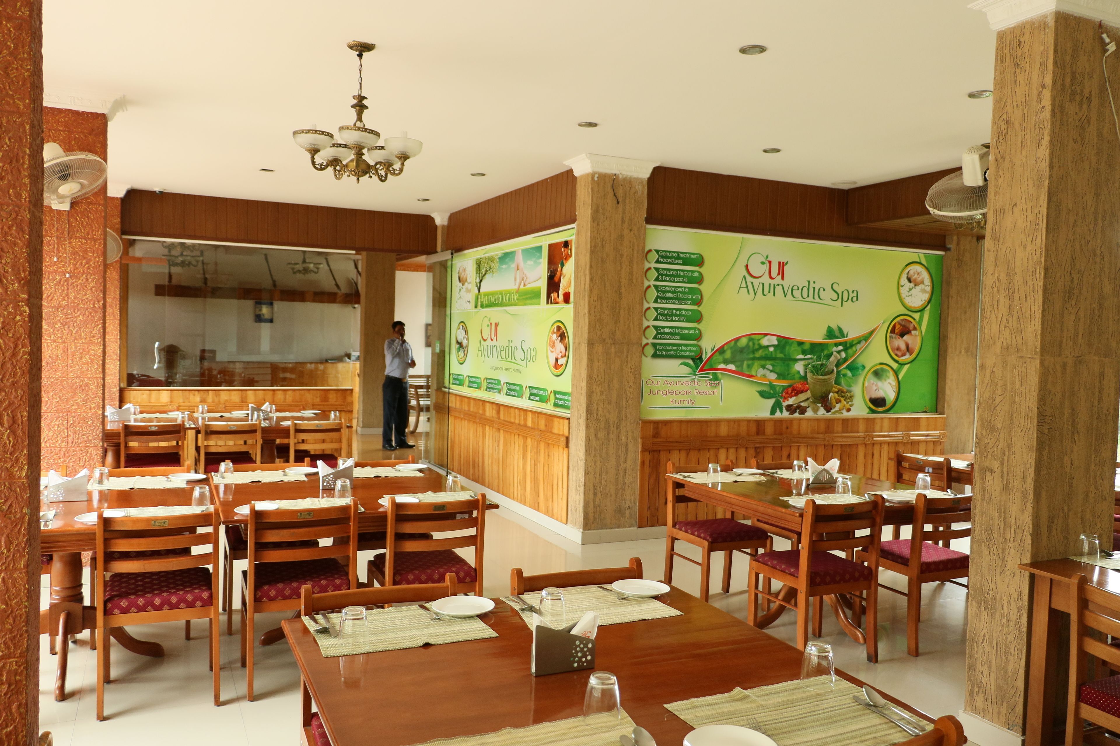 Un restaurante con mesas y sillas de madera, un mural grande que anuncia servicios de Spa Ayurveda, y una persona al fondo.