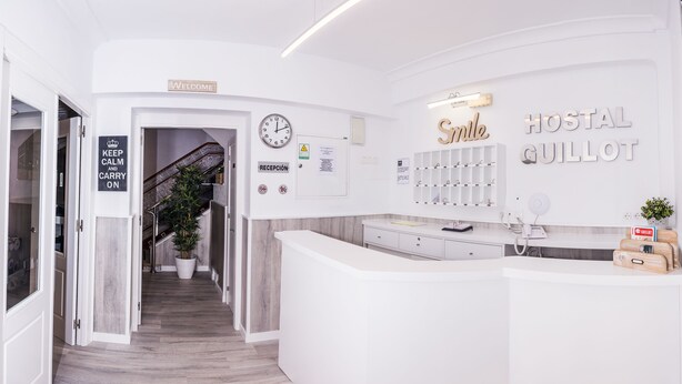 A reception area with a clock, a welcome sign, a reception desk, and a wall with 'Smile' and 'HOSTAL GUILLOT' signs. - Hostal Guillot (Torremolinos)