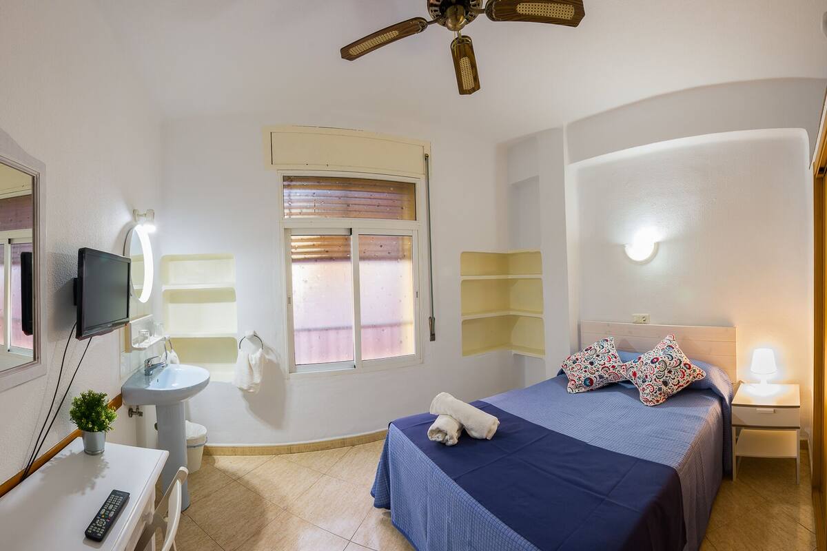 Habitación de hotel con cama, escritorio con silla, baño con lavamanos y espejo, ventilador de techo y ventana con persianas.