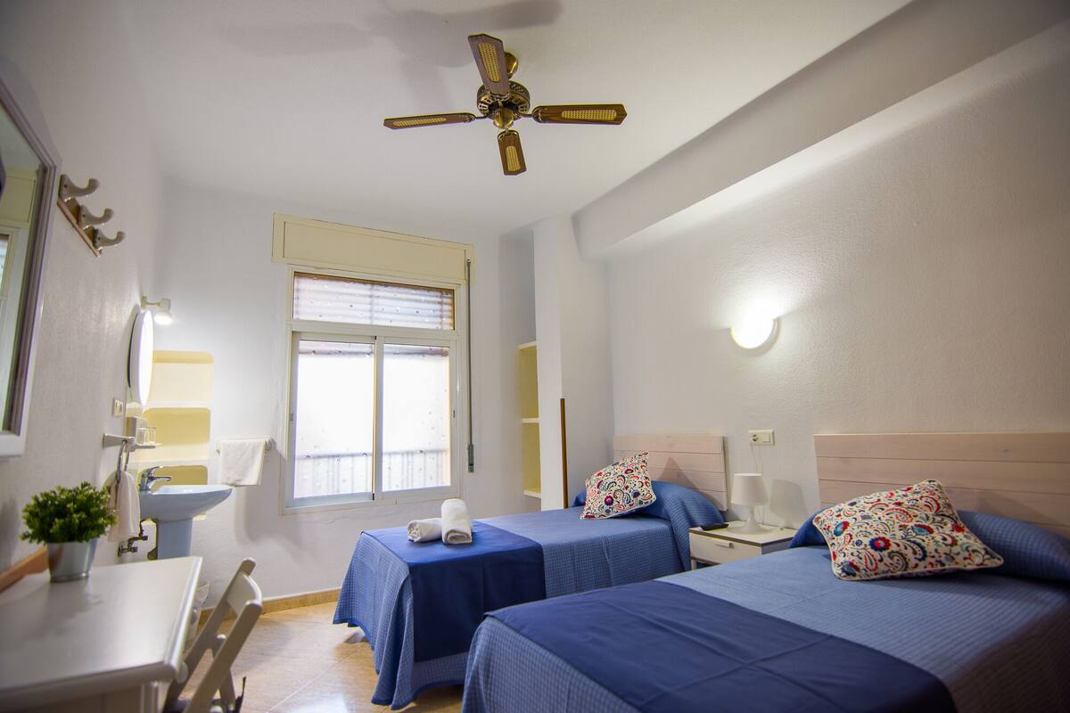 Habitación de hotel con dos camas, un ventilador de techo, una ventana con persianas, un lavabo y una pequeña mesa con una planta.