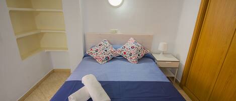 Un lit bien fait, recouvert d’une couvre-lit bleue et agrémenté de deux coussins décoratifs, une lampe de chevet blanche et une porte en bois.
