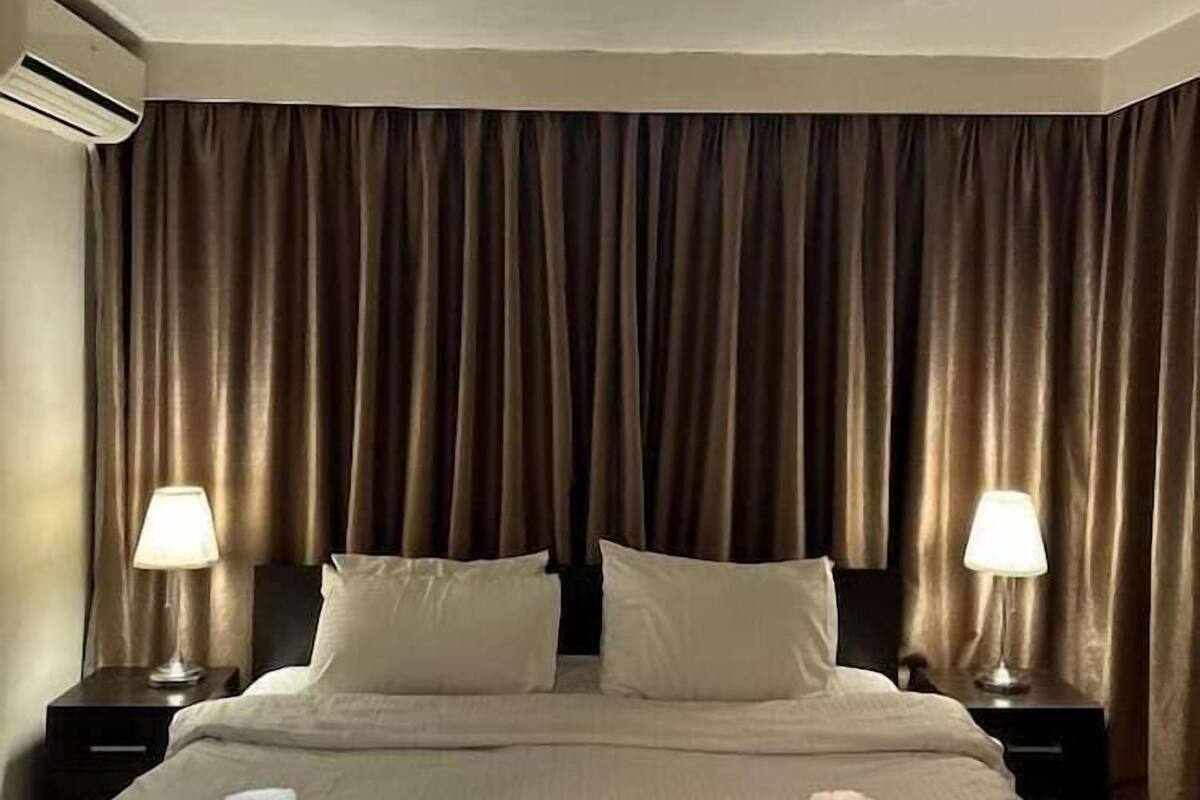 A hotel room with a bed, two bedside lamps, a ceiling light, and a curtain.