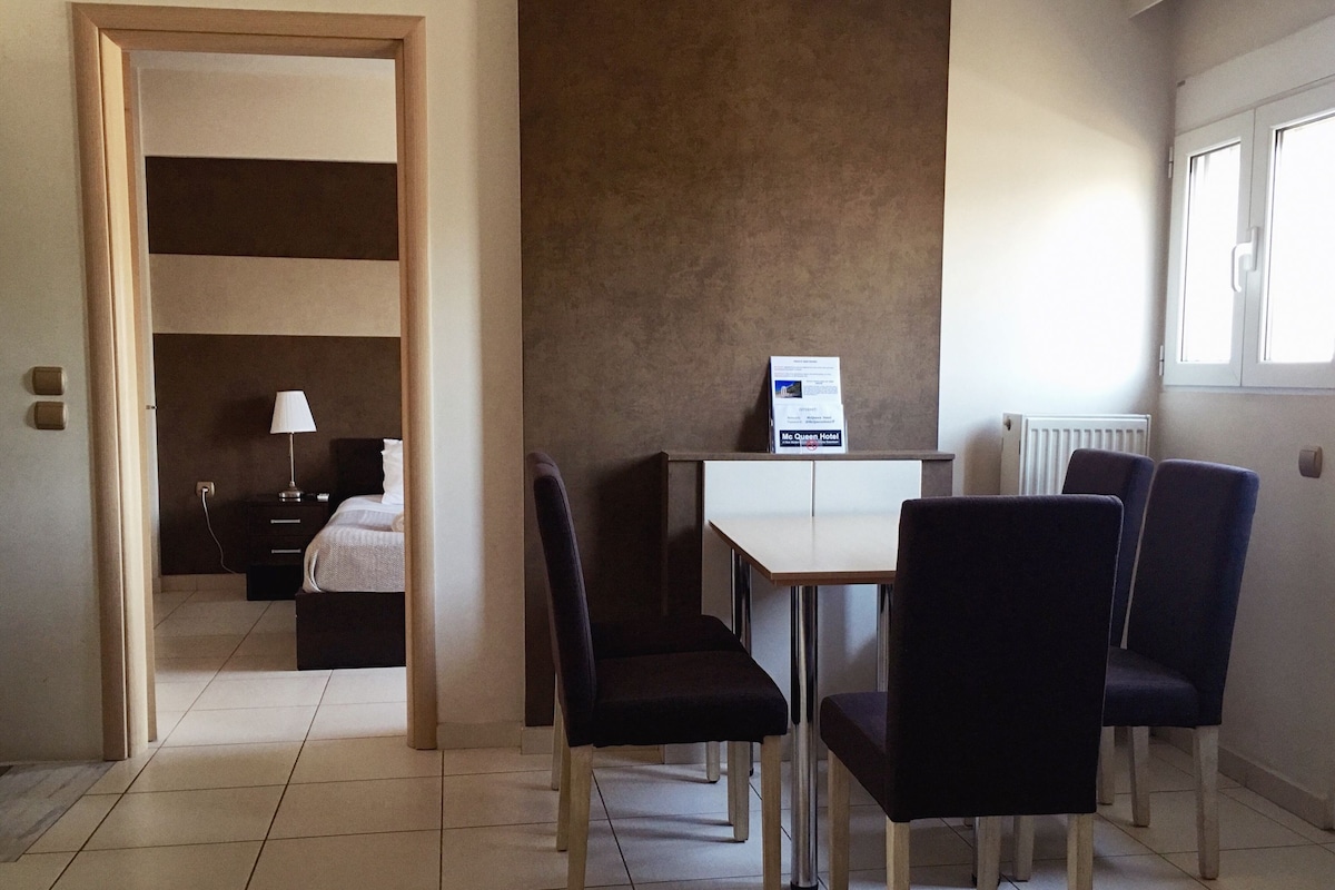 A hotel room with a bed, a desk, and two chairs. There is a sign on the desk and a window with blinds.