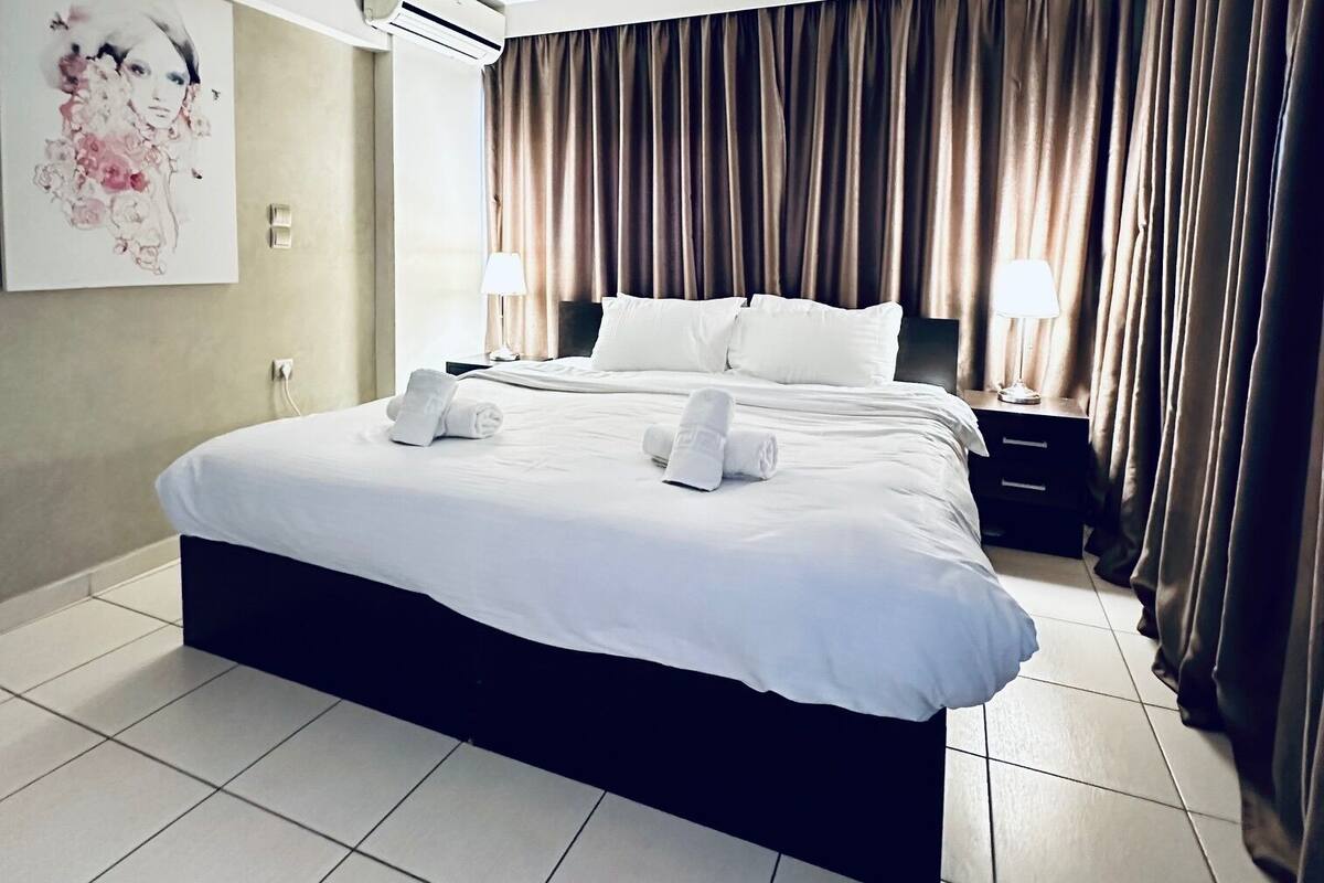 A hotel room with a large bed, a ceiling fan, an air conditioning unit, a bedside table with a lamp, and a painting on the wall.