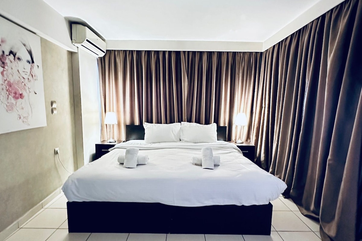 A modern hotel room with a large bed, a ceiling fan, and a view of the city through the window.