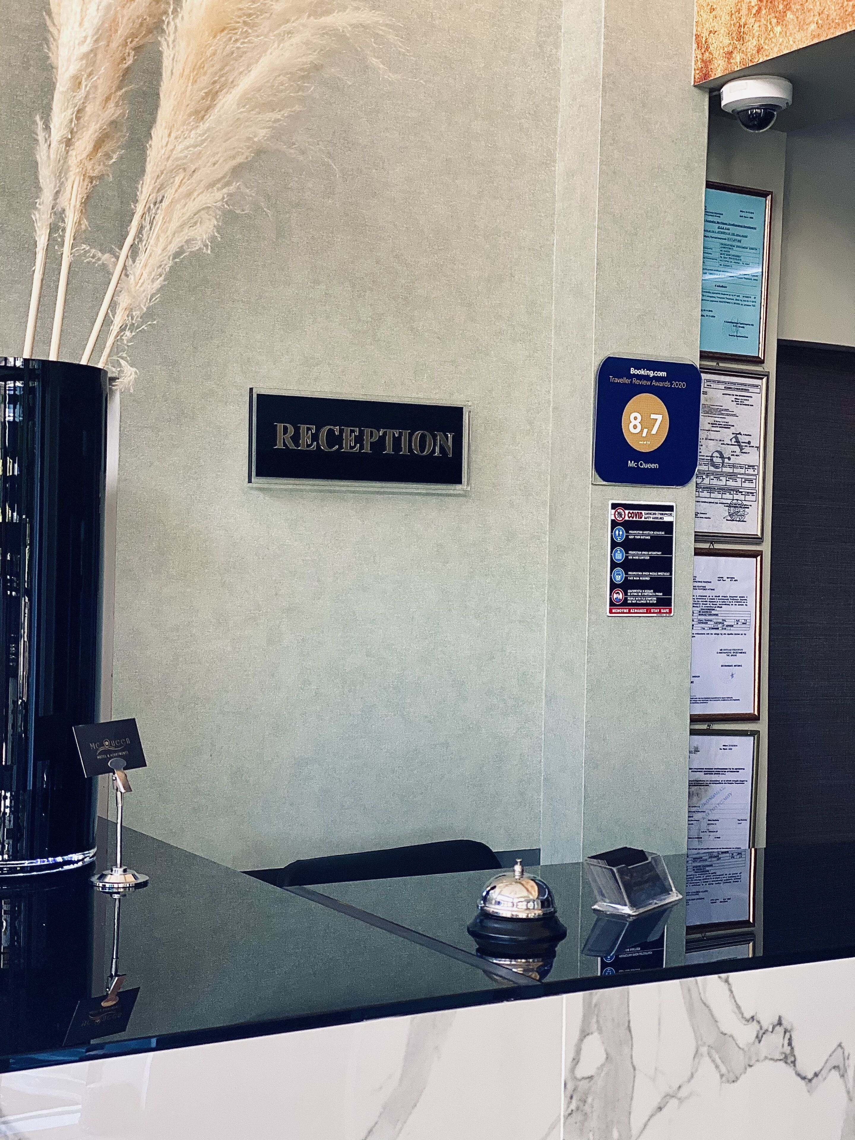 Een receptiebalie met een bord waarop 'RECEPTION' staat, een 'Booking.com' prijsplaquette, veiligheidsaandachtspunten voor COVID-19 en een 'McQueen' plaquette.