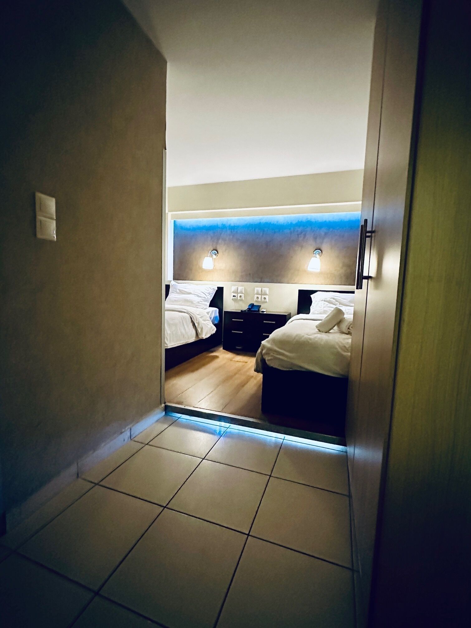 Habitación de hotel con dos camas, piso de madera, pared de acento azul y una mesita de noche con lámpara.