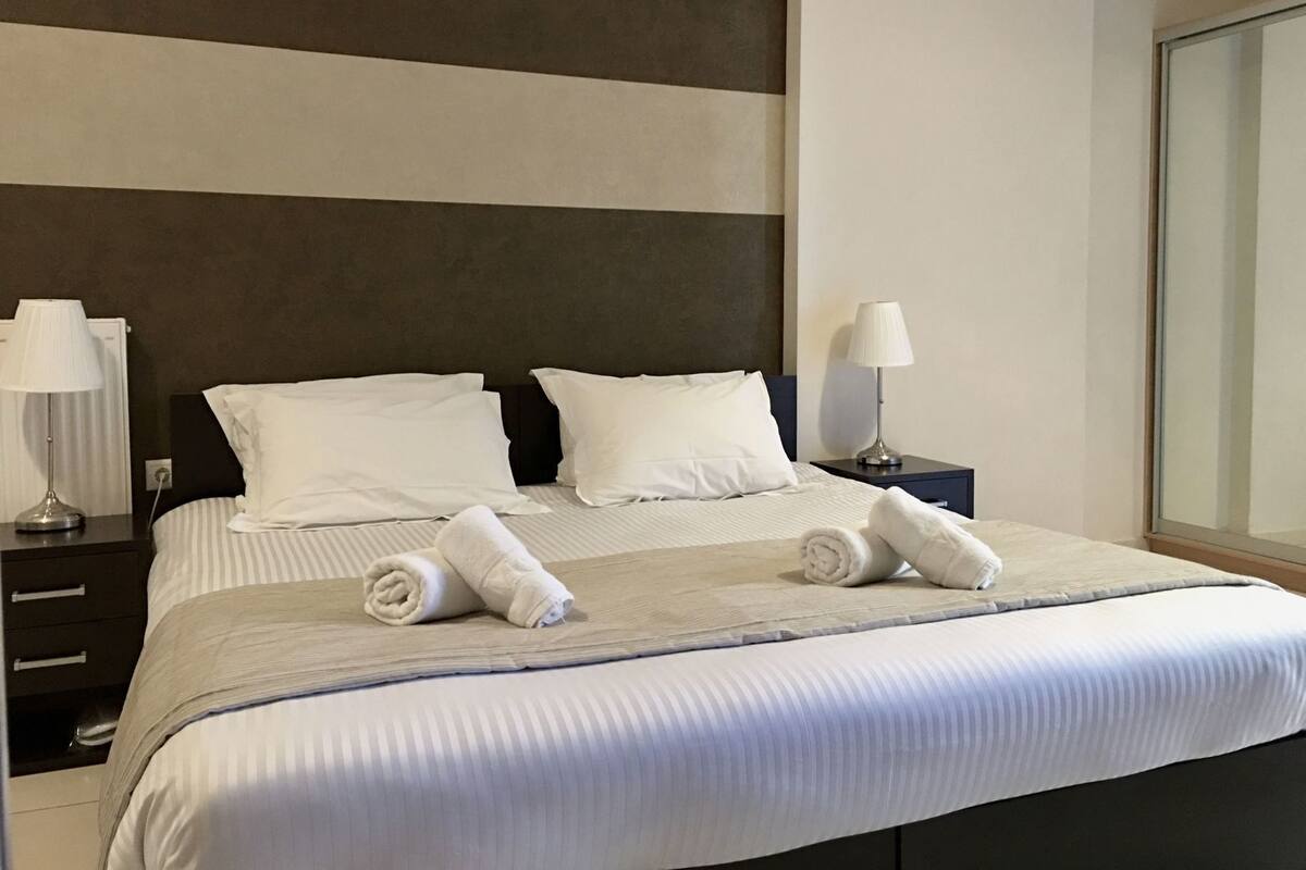 A neatly made bed with white linens, two pillows, and two rolled towels. There is a nightstand with a lamp on each side of the bed.