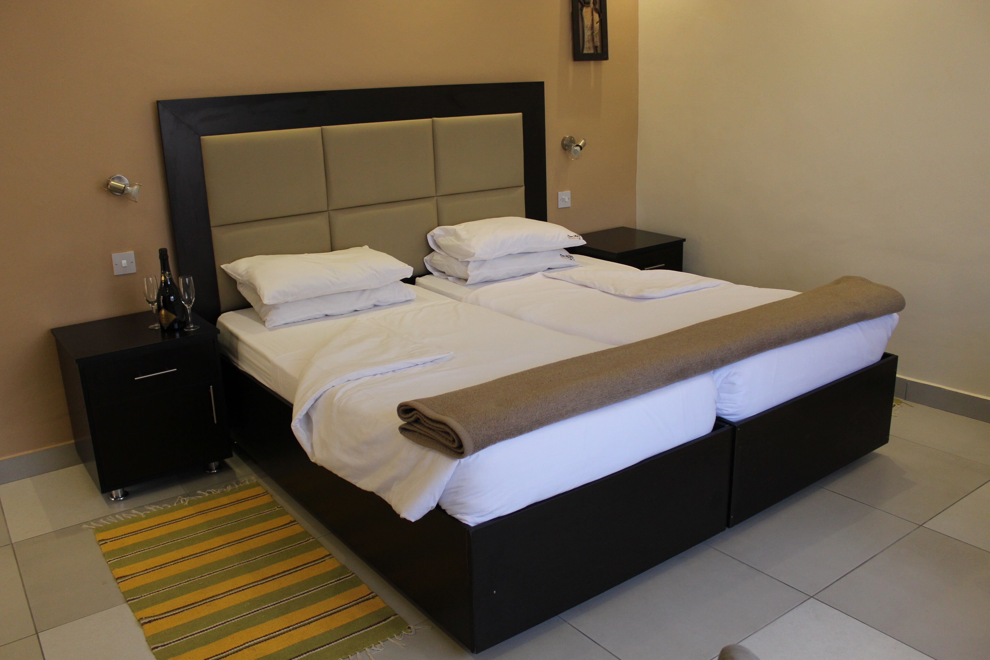 A neatly made bed with white linens and a brown throw blanket, a bedside table with a bottle and glasses, and a striped rug on a tiled floor.