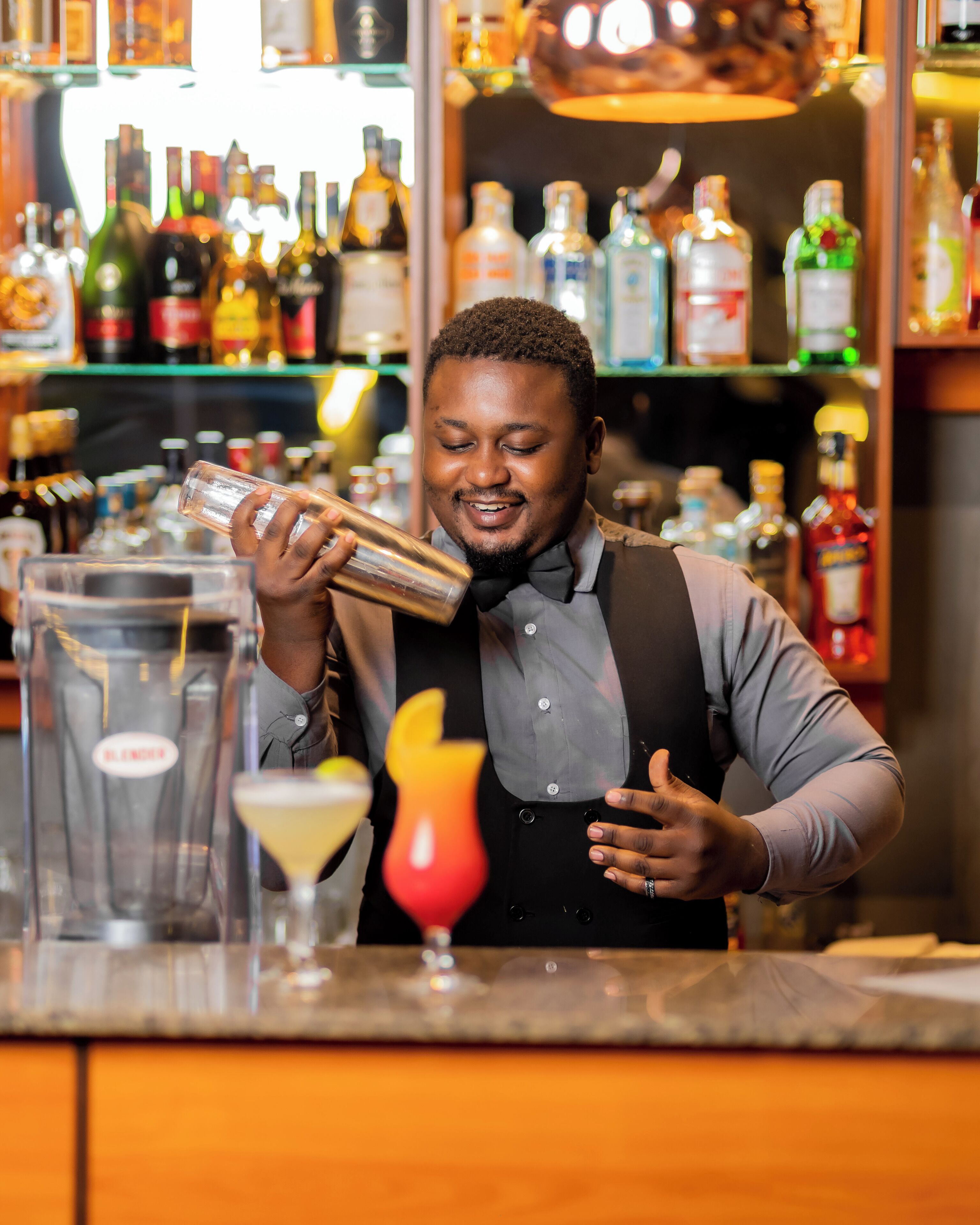 A bartender preparing a drink behind a bar with various bottles and a cocktail shaker.