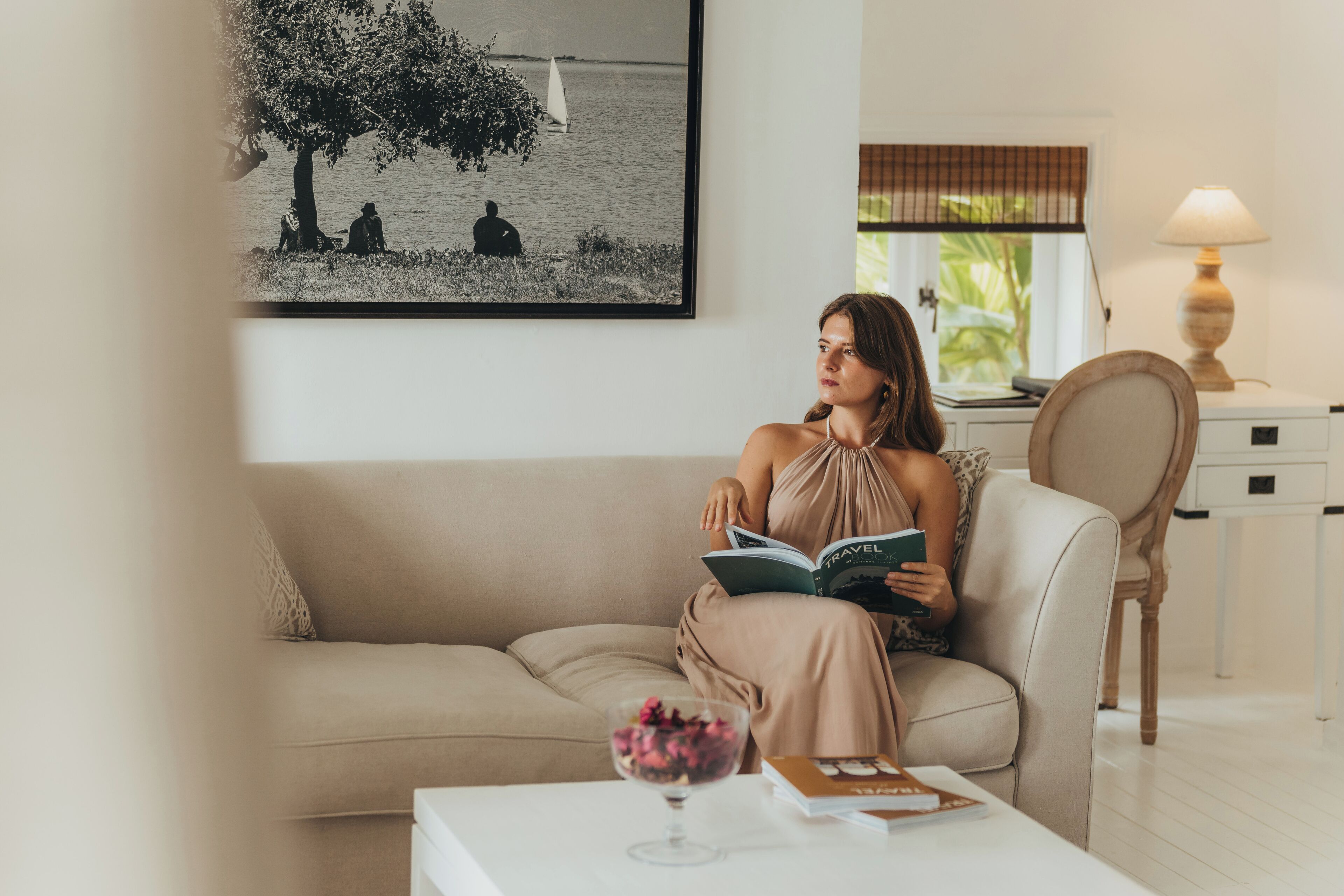 Una mujer sentada en un sofá beige leyendo un libro, con una foto blanco y negro enmarcada de personas junto al agua en la pared detrás de ella.