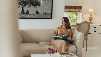 Una mujer sentada en un sofá beige leyendo un libro, con una foto blanco y negro enmarcada de personas junto al agua en la pared detrás de ella.