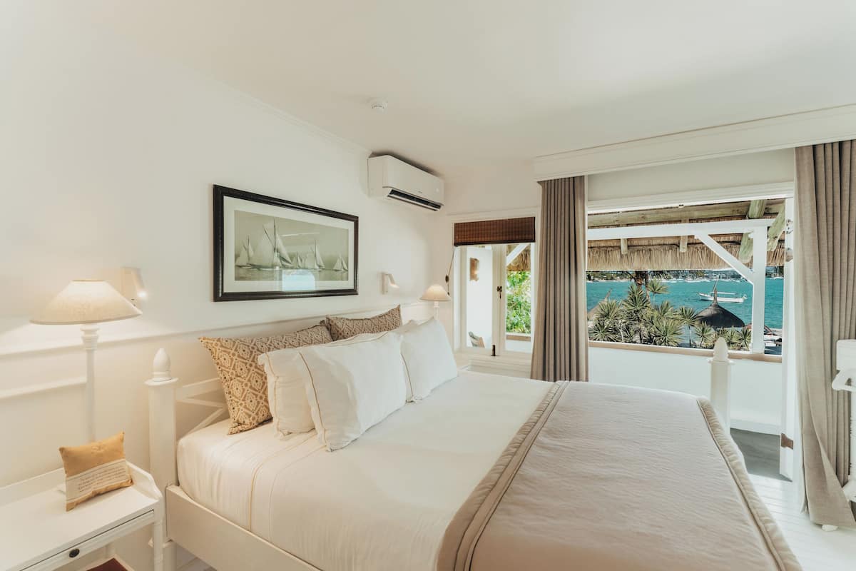 A hotel room with a large bed, a framed picture on the wall, an air conditioning unit, and a view of the sea through the window.