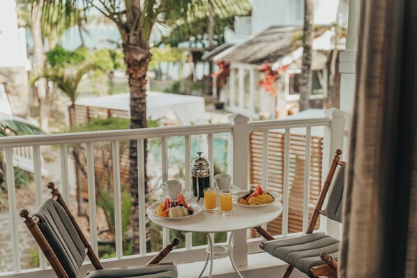 Una mesa blanca con alimentos para el desayuno, una cafetera y dos sillas en un balcón con vistas a palmeras y un edificio.