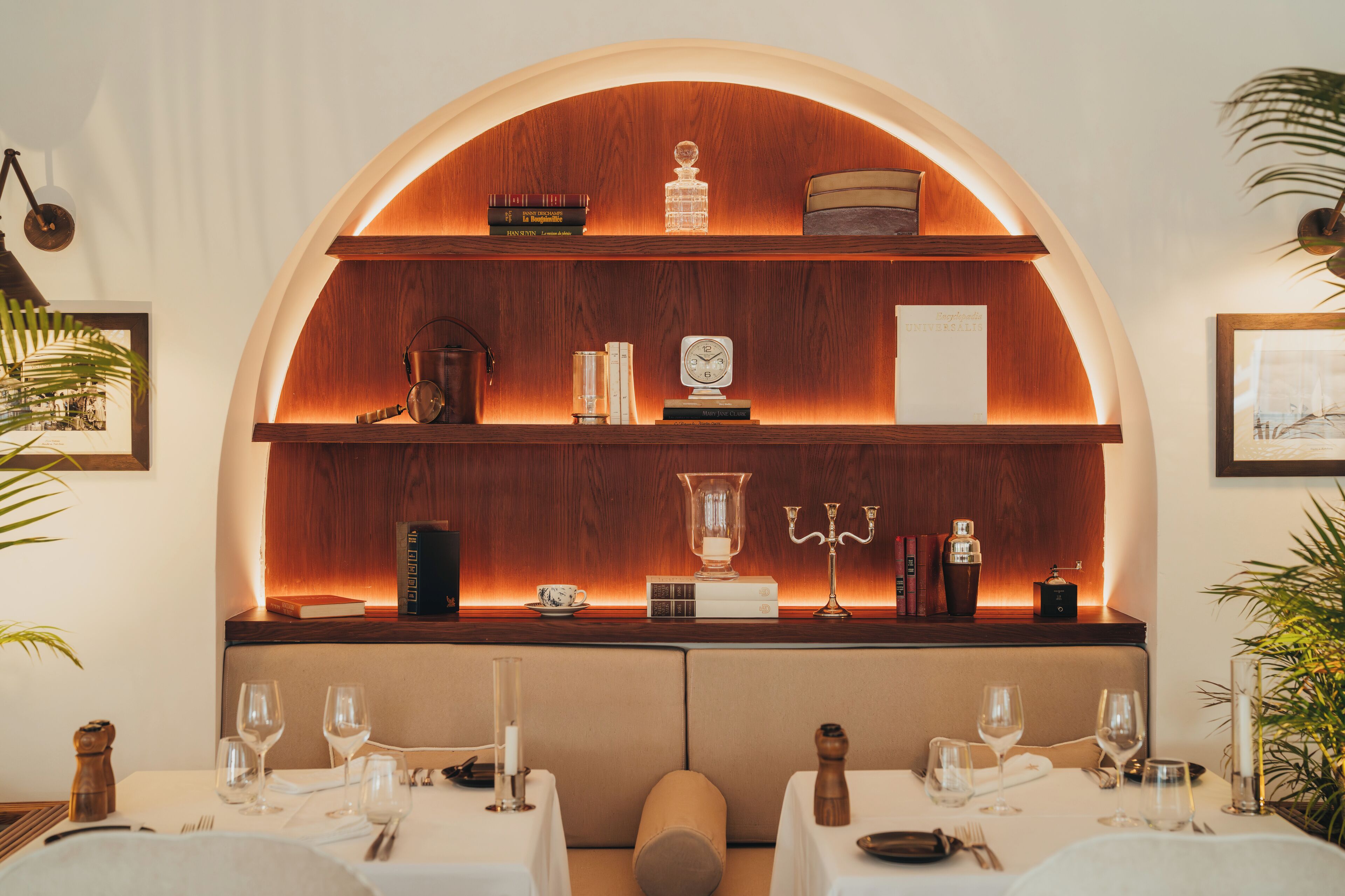Un comedor con un estante de madera que muestra libros, un reloj y un candelabro. Las mesas están dispuestas con copas de vino y cubiertos.