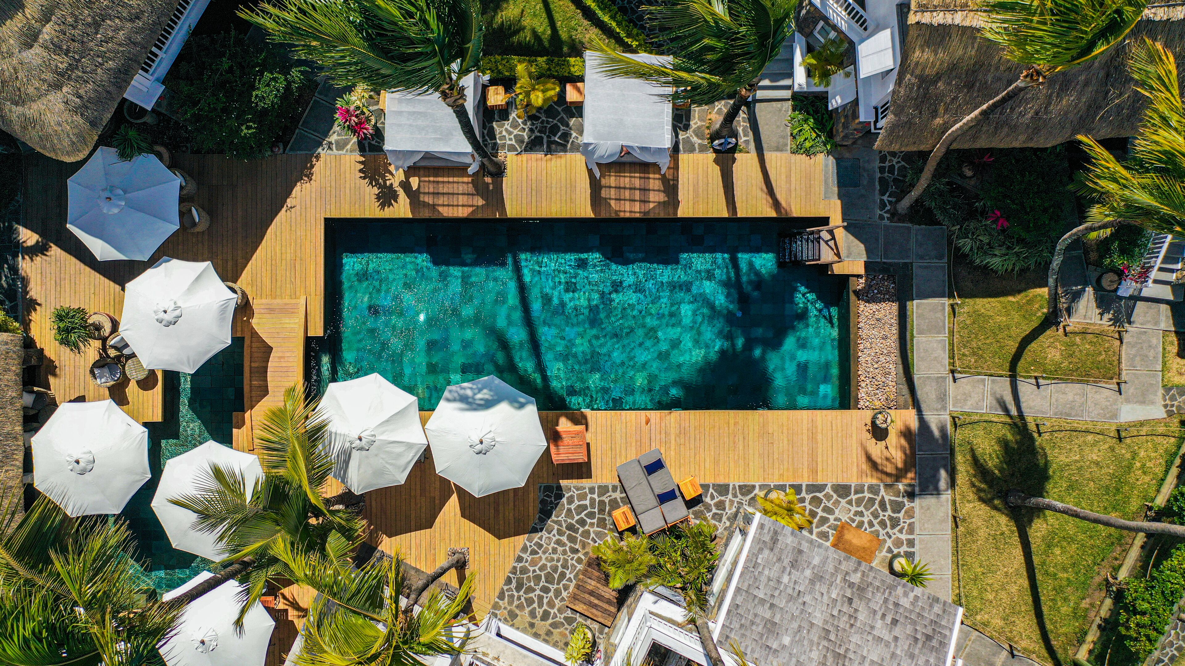 An aerial view of a resort with a rectangular pool surrounded by wooden decking, white umbrellas, and lounge chairs.