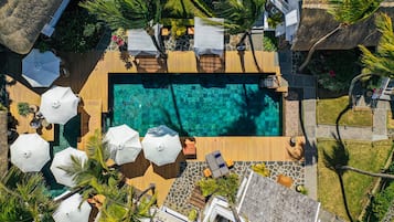 An aerial view of a resort with a rectangular pool surrounded by wooden decking, white umbrellas, and lounge chairs.