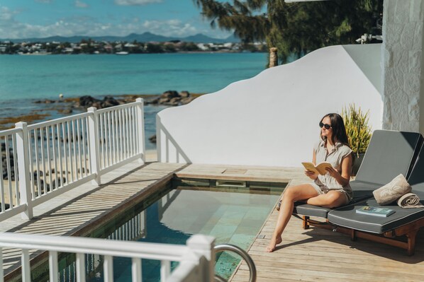 Una mujer sentada en una silla de descanso junto a una piscina, con vistas al mar y montañas al fondo.