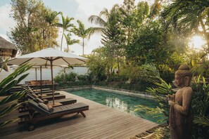 Una terraza de madera con sillones de descanso y una piscina, rodeada de exuberante vegetación y una estatua de Buda.