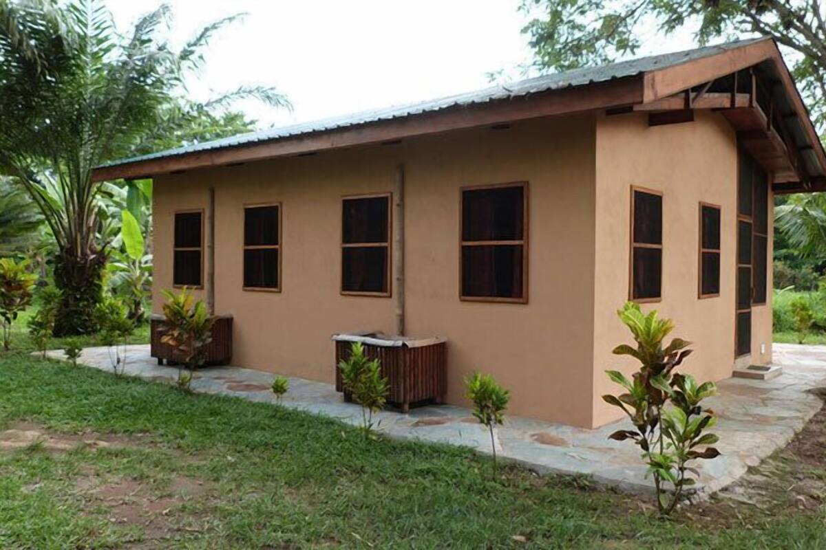 A single-story building with a flat roof and a simple, functional design. It features a series of windows aligned along the side, a small porch area with a bench, and is surrounded by a well-maintained garden with various plants and a stone pathway leading to the entrance.