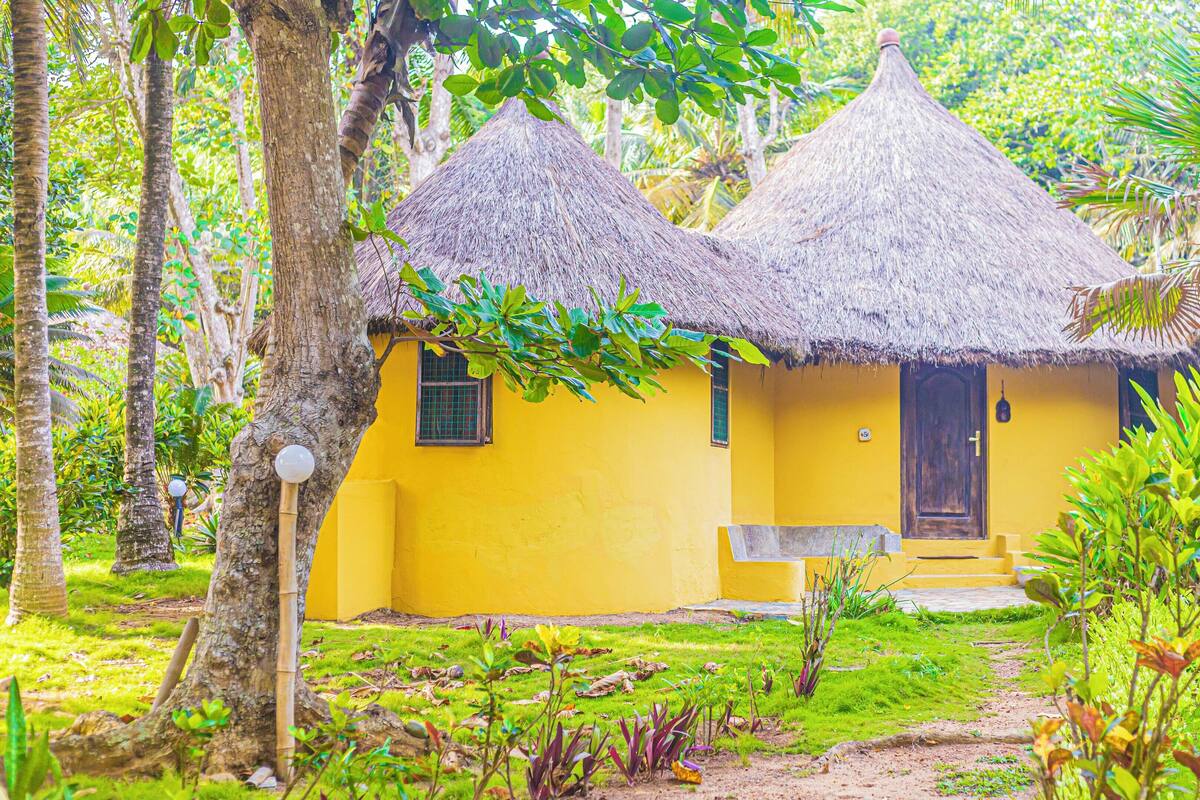 A yellow building with thatched roofs, surrounded by lush greenery.