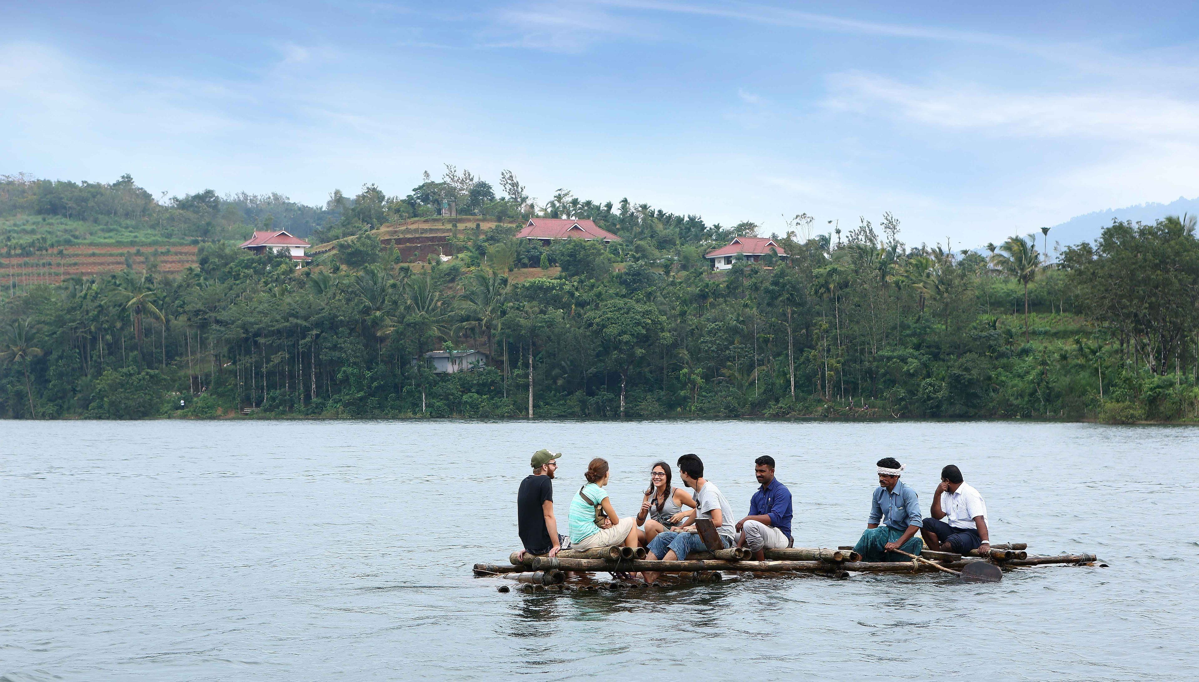 Un groupe de personnes se trouve sur un radeau en bambou, sur un lac paisible, avec des maisons et de la végétation en arrière-plan.