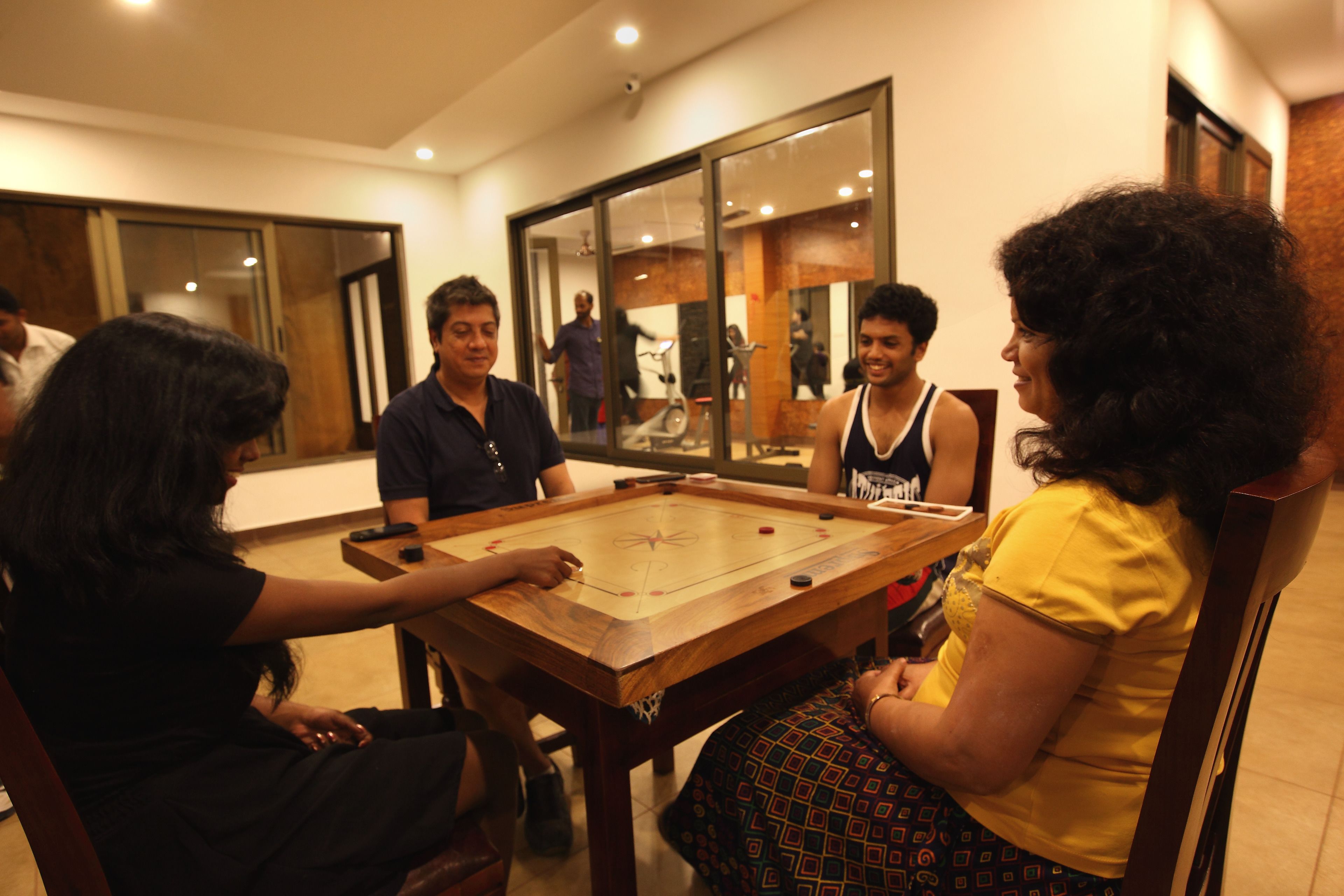 Un grupo de personas jugando un juego de mesa en una mesa de madera, en una habitación con un espejo grande y un ventilador de techo.