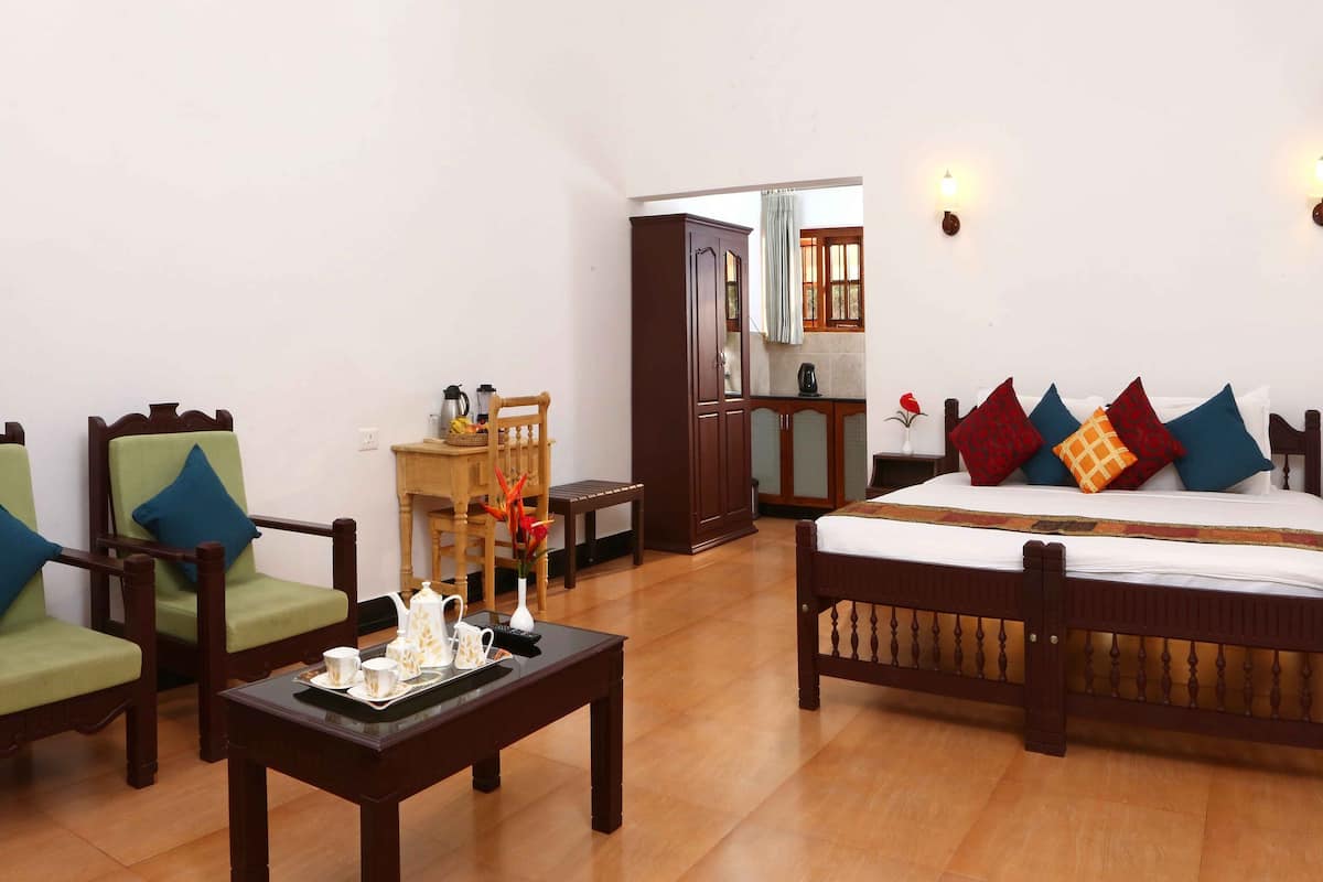 Ein Hotelzimmer mit einem hölzernen Bett, zwei grünen Stühlen, einem kleinen Tisch mit Frühstücksgebäck und Blick in die Küche durch eine geöffnete Tür.