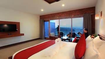 Habitación de hotel con una cama grande, televisor montado en la pared, balcón con vistas y zona de comedor con mesa y sillas.