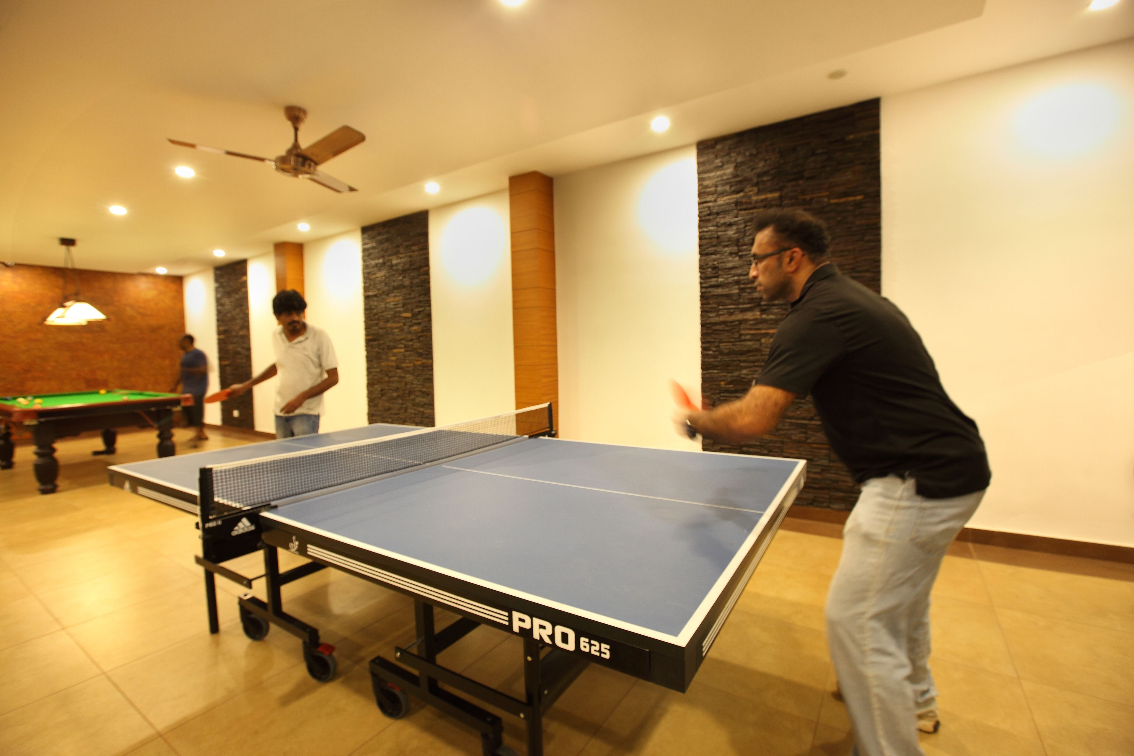 A ping pong table in a room with a ceiling fan, a billiards table, and a person playing ping pong.