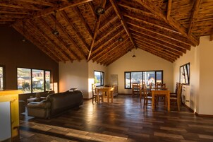 Un amplio salón con techo de madera alta, un sofá, una mesa de comedor con sillas y ventanas grandes que ofrecen vistas al exterior.