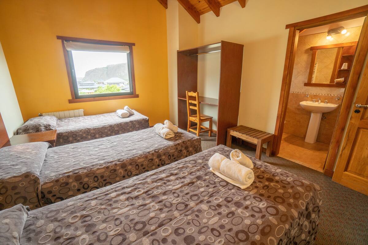 Una habitación con dos camas, una ventana, una silla de madera, un banco y un baño con lavabo y espejo.
