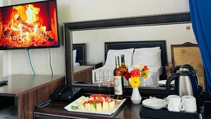 Ein Hotelzimmer mit einem Bett, einem Schreibtisch mit Obstteller, einer Kaffeemaschine und einem Fernseher, der einen Kamin zeigt.