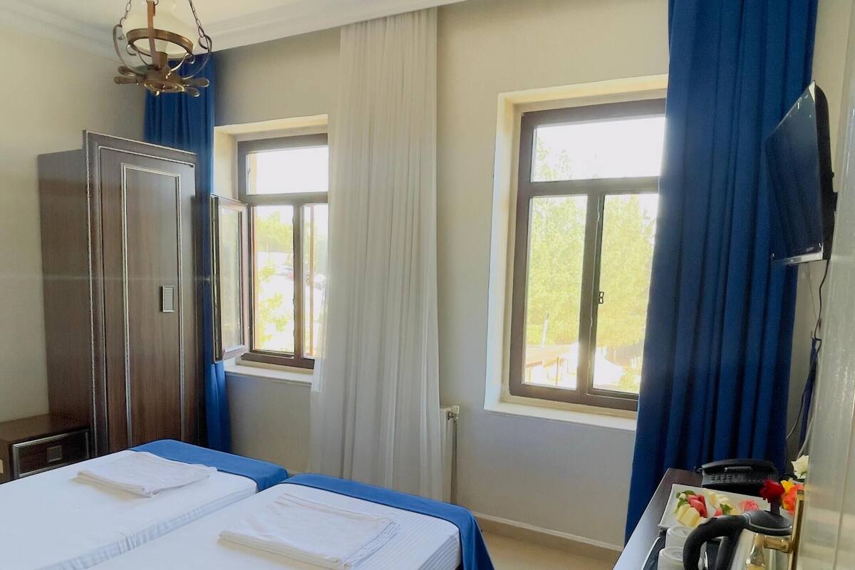 Hotellihuone, jossa on suuri sänky, vaatekaappi, kirjoituspöytä, jossa on puhelin ja vedenkeitin, sekä kaksi ikkunaa, joissa on verhot.