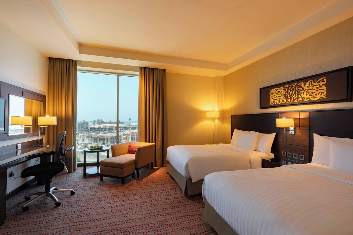A hotel room with two beds, a desk, a chair, and a view of the city.