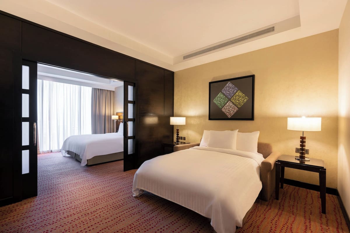 A hotel room with a large bed, a bedside table with a lamp, a wall-mounted artwork, and a view through an open door.