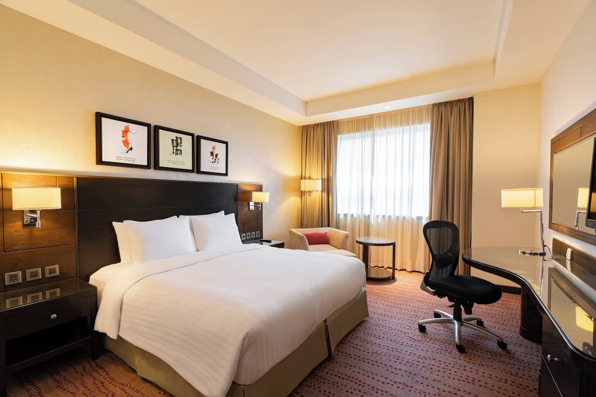 A hotel room with a large bed, a desk, a chair, and a TV. There are two framed pictures on the wall and a window with curtains.