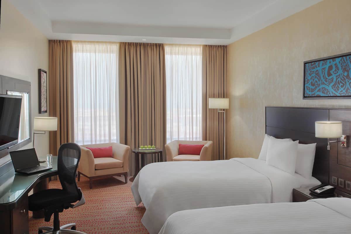 A hotel room with two beds, a desk with a laptop, a TV, and a small table with a lamp.