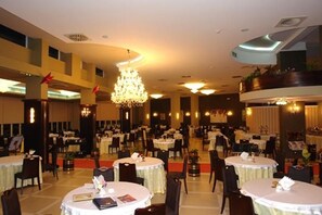 A spacious dining area with multiple round tables and chairs, a central chandelier, and a high ceiling with recessed lighting. - Bleart (Rrashbull)