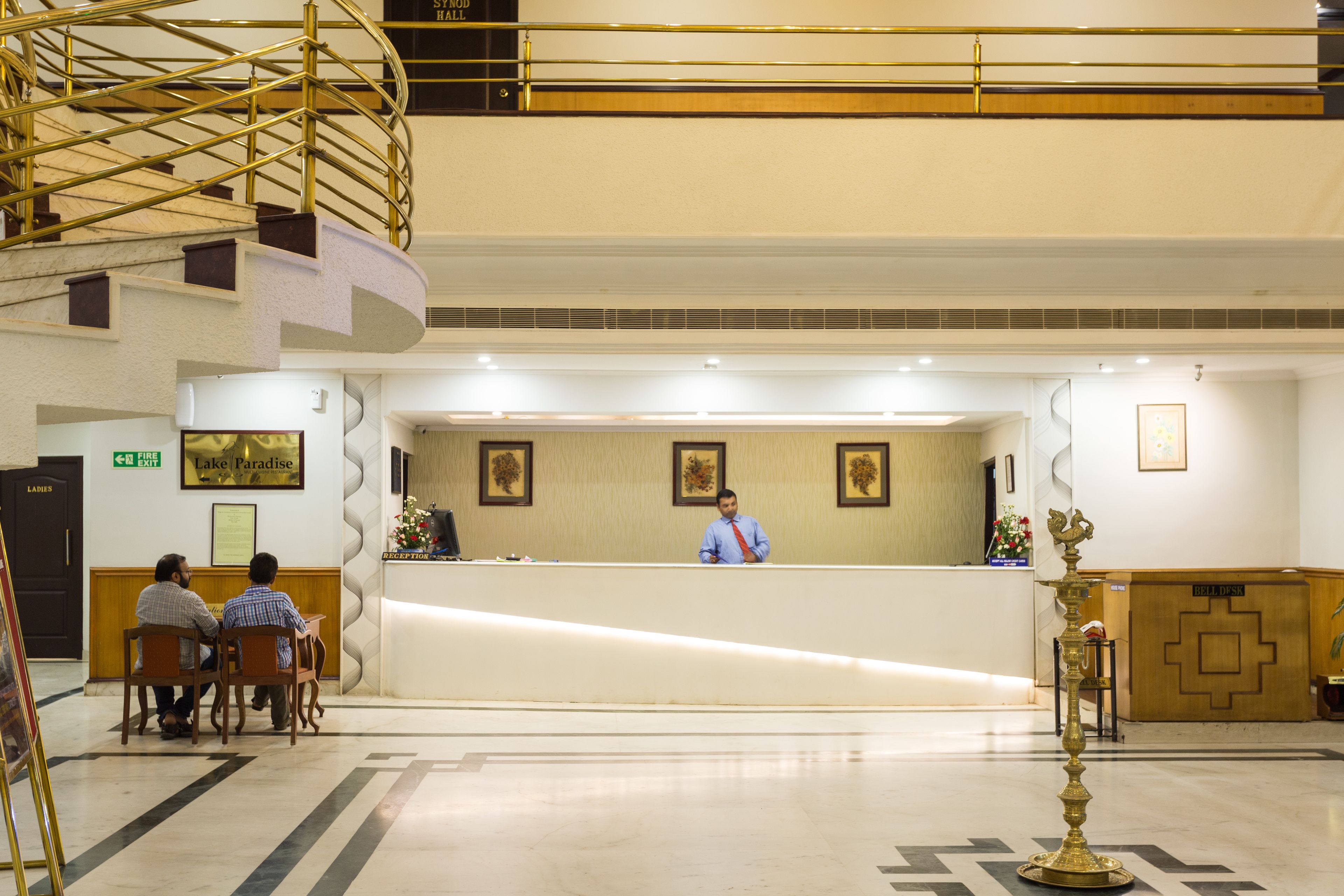 A hotel reception area with a receptionist, two seated guests, a staircase, and a reception desk.