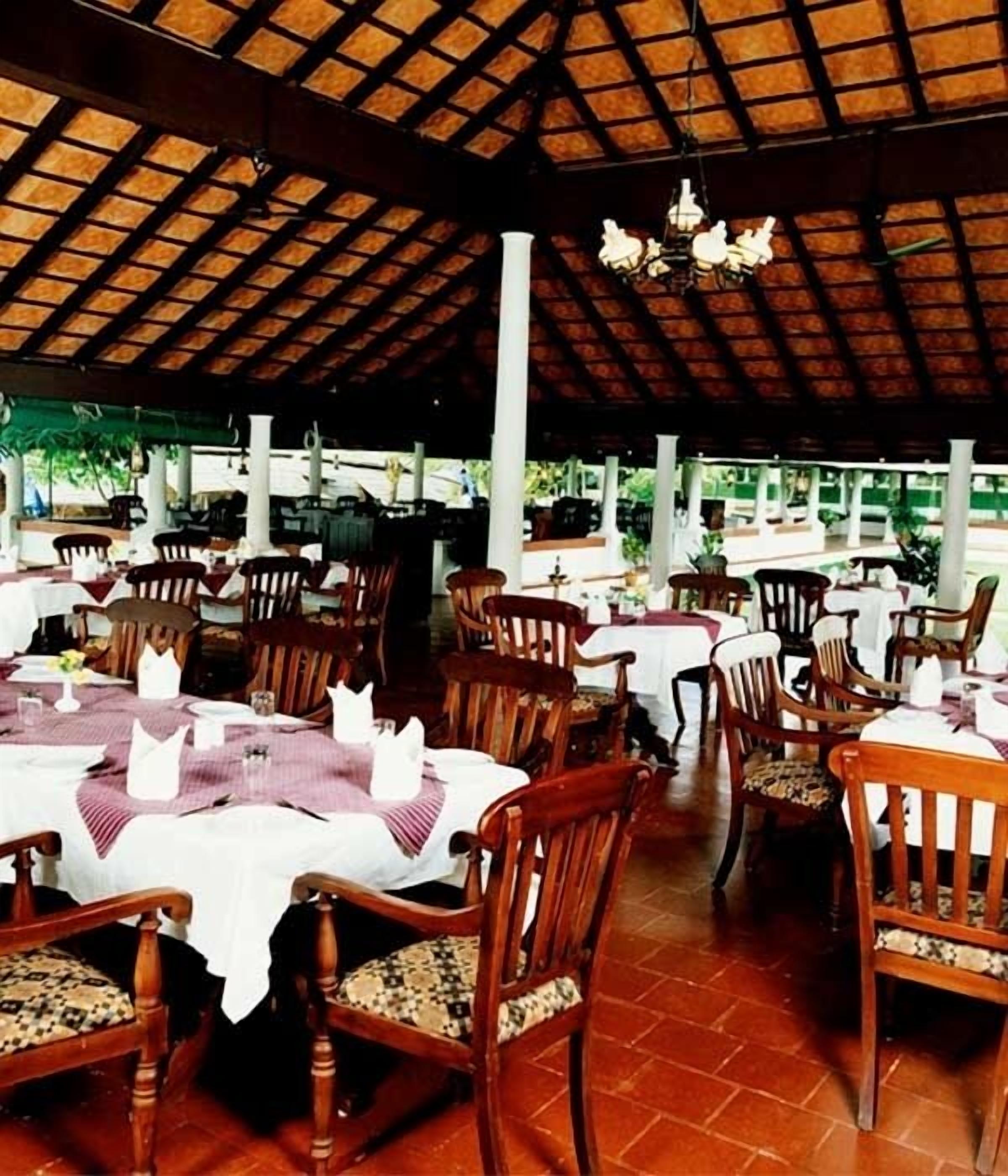 An outdoor restaurant with wooden tables and chairs, covered by a thatched roof.