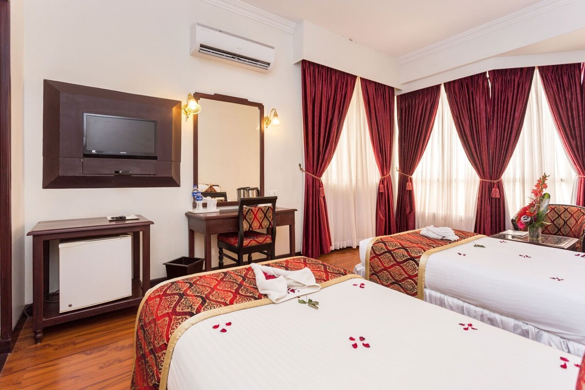 A hotel room with two beds, a desk, a chair, a television, and a large window with red curtains.