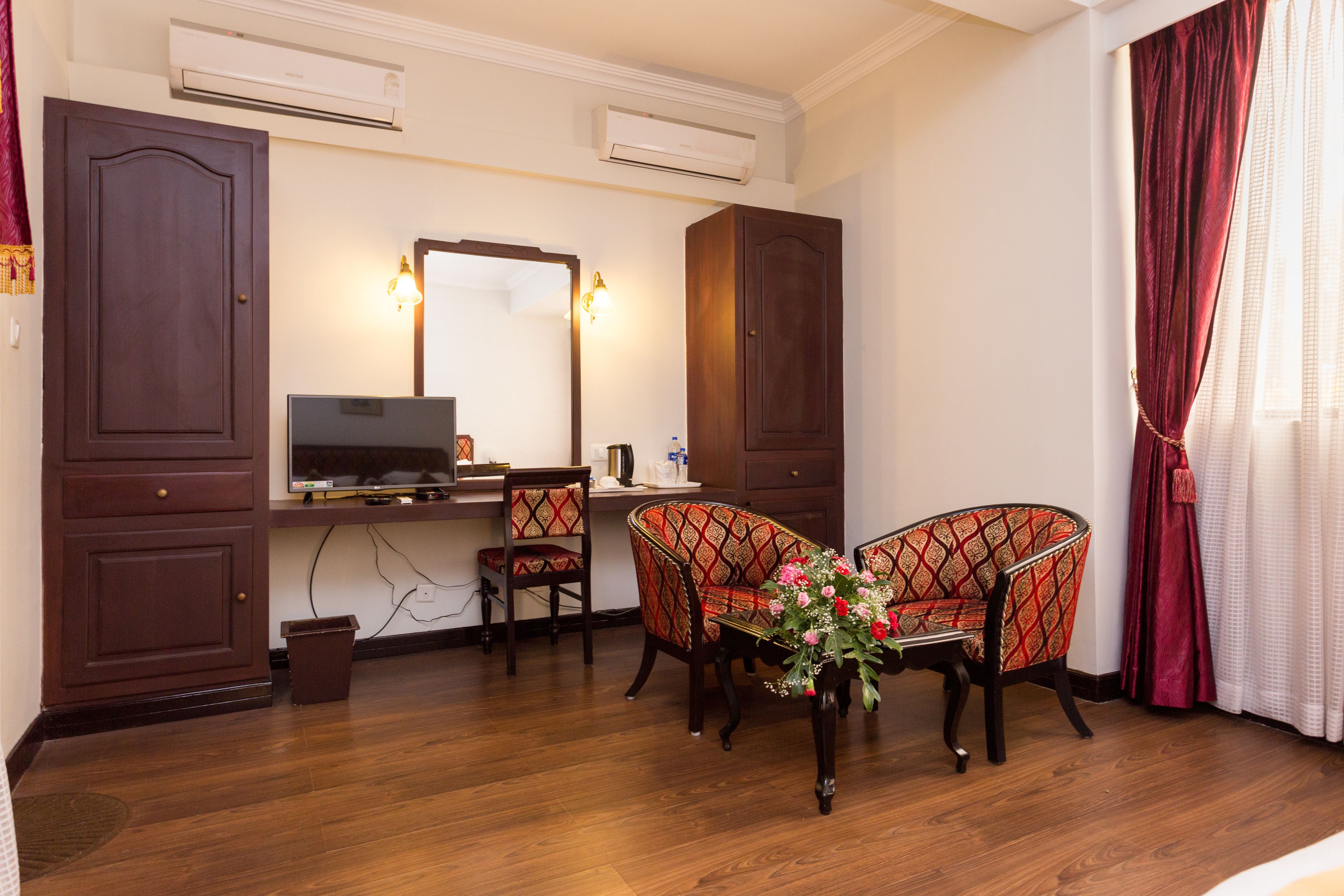 A hotel room with a wooden desk, two patterned chairs, a mirror, and a red curtain.